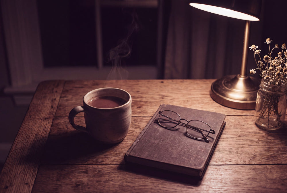 A kitchen table at 2am: a mug of cocoa, reading glasses on a closed notebook, a small lamp and a jar of chamomile.